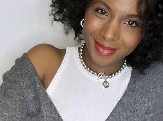 Jacqueline is smiling at the camera. Her hair is black and very curly and she is wearing a white top and a grey jumper.