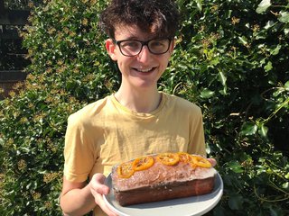 Jamie, in a yellow T-shirt with an orange cake he baked in front of a hedge.