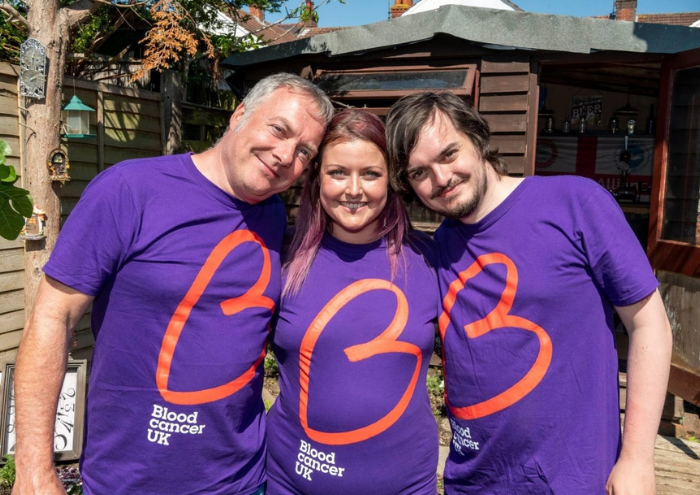 A woman - Jemma - shares a moment with two men. They all wear purple Blood Cancer UK T shirts.