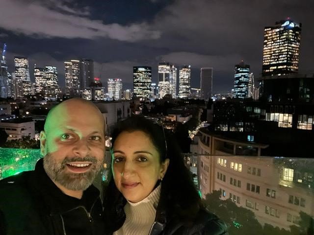Kal (on the right) with her husband. They are standing together on a glass balcony with a view of a city at night behind them.