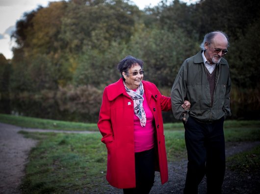 A stock image of a man and a woman walking arm in arm through a dimly lit park, smiling slightly and wearing jackets.