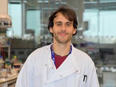 A man with dark hair in a lab, wearing a white lab coat smiling at the camera.