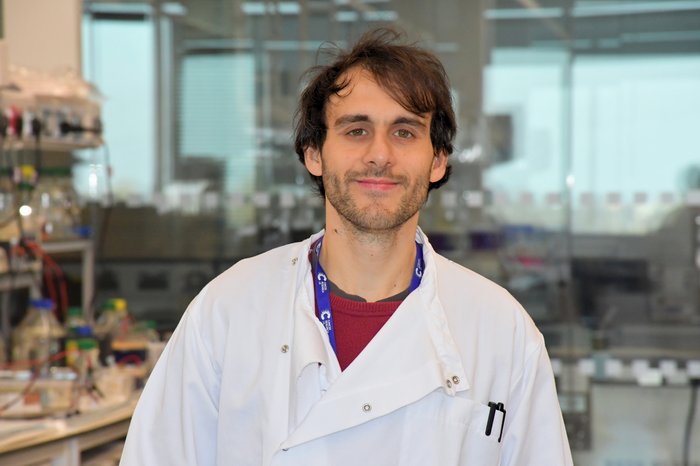 A man with dark hair in a lab, wearing a white lab coat smiling at the camera.