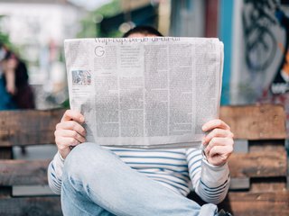 A stock image of a man sitting on a bench in the street, holding a newspaper up over his head.