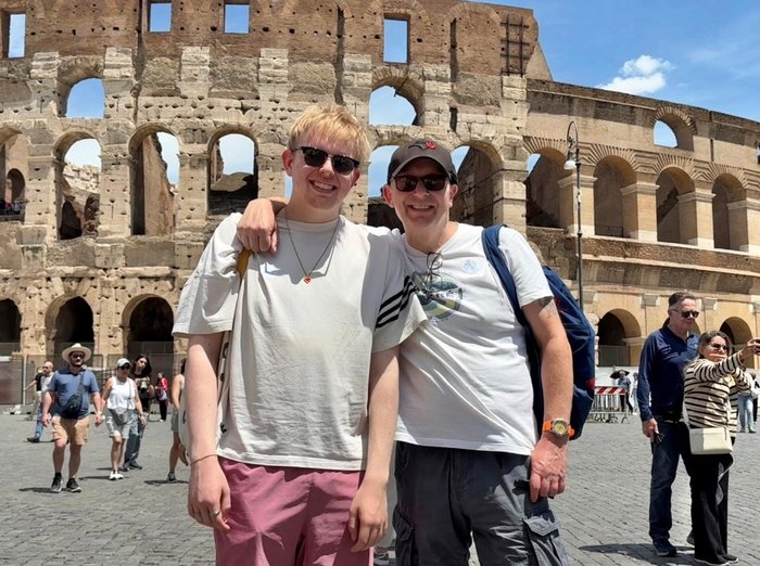 Mark stands in front of the colosseum in Rome facing the camera. He has a hat and sunglasses on. His teenage son stands next to him. They are both smiling.