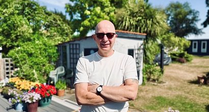 Mark stands facing the camera in his garden. It's sunny and he is smiling. There is a shed, lawns and flowerbeds in the background.