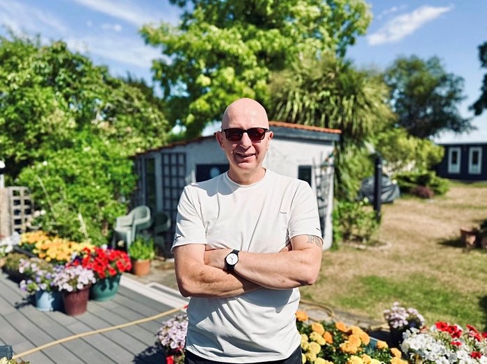 Mark stands facing the camera in his garden. It's sunny and he is smiling. There is a shed, lawns and flowerbeds in the background.