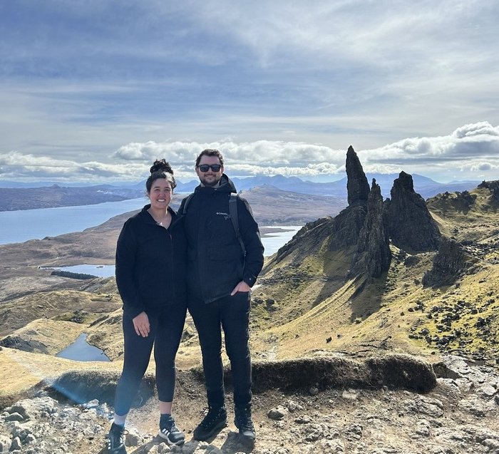 Matt and his partner, standing together on top of a mountain or large hill, with a view of hills, sky and water behind them.