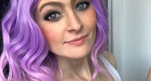 A close up of Megan Rhodes wearing a bright purple wavey wig, looking straight at the camera.