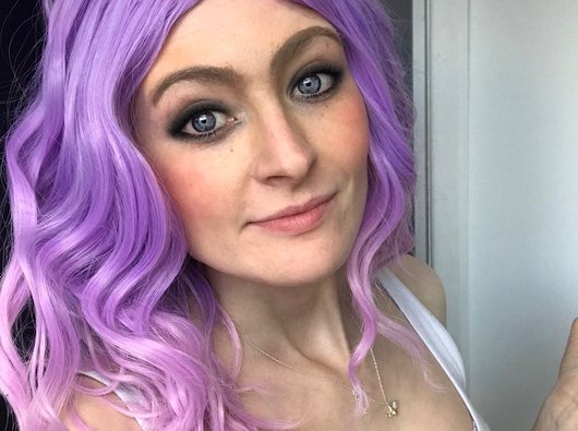 A close up of Megan Rhodes wearing a bright purple wavey wig, looking straight at the camera.