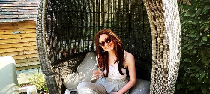 Megan Rhodes smiling, wearing sunglasses and holding a drink, sitting in an egg chair outside in the sun.