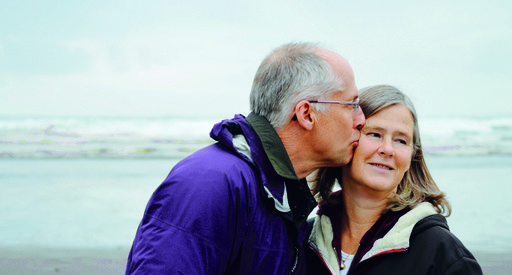 A middle aged couple on a beach in coats. Man kissing woman.