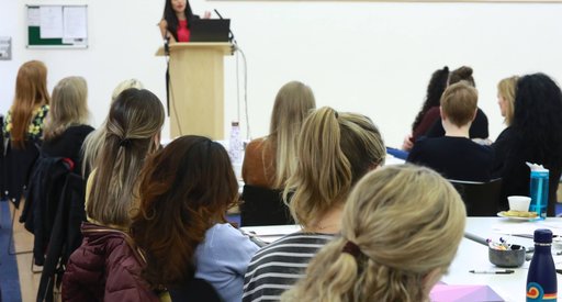 At a meeting, a woman stands and speaks with a laptop while an all-female seated crowd listens.