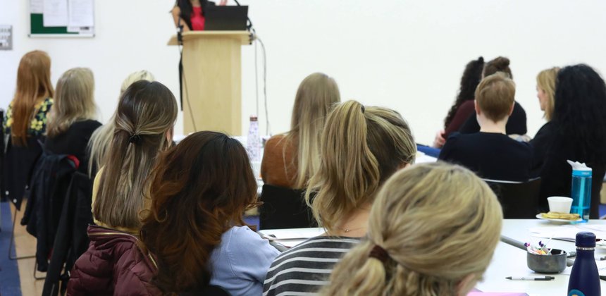 At a meeting, a woman stands and speaks with a laptop while an all-female seated crowd listens.