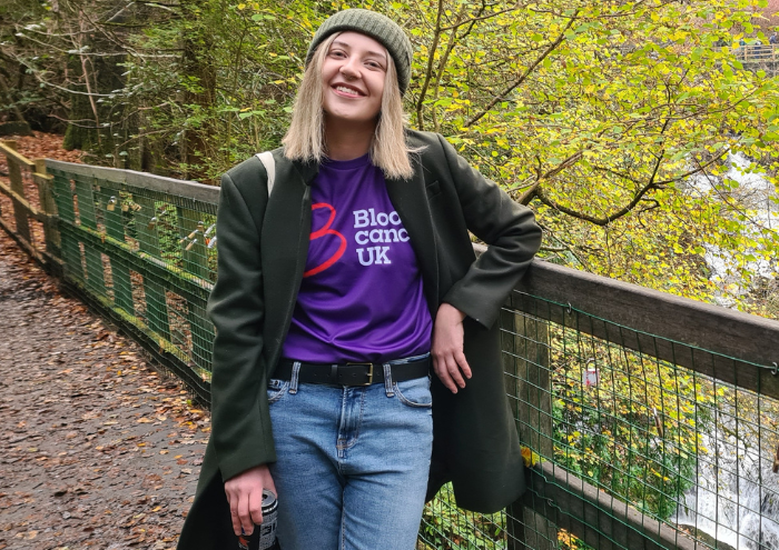 A young woman - Nichola - wearing a Blood Cancer UK t shirt under a winter coat stands smiling on a bridge in the countryside.