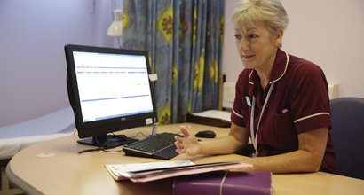 A nurse sits at her desk in a hospital with a computer and notepads.