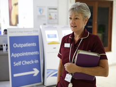 A nurse, holding a folder, stands in a hospital waiting room with a receptionist behind her.