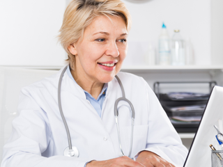 A nurse, in a lab coat and with a stethoscope around her neck, sits at a desk typing on a laptop