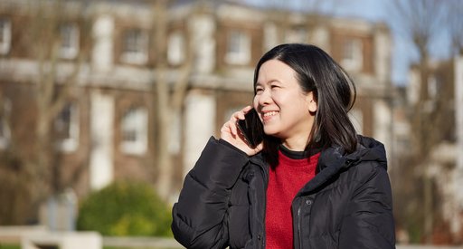 A woman on a telephone call stands in a park with a large house blurred in the background.