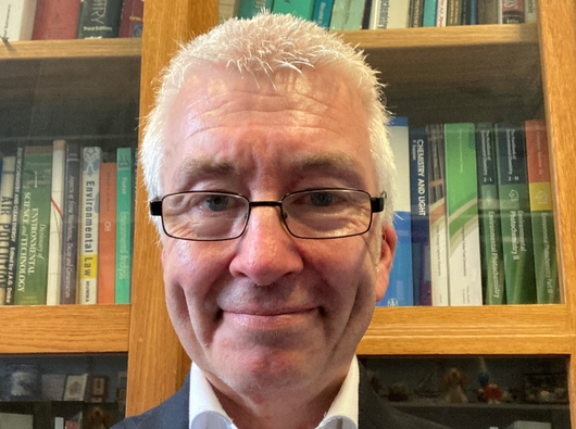 Photo of Peter, smiling and wearing a blazer standing in front of his book shelf.