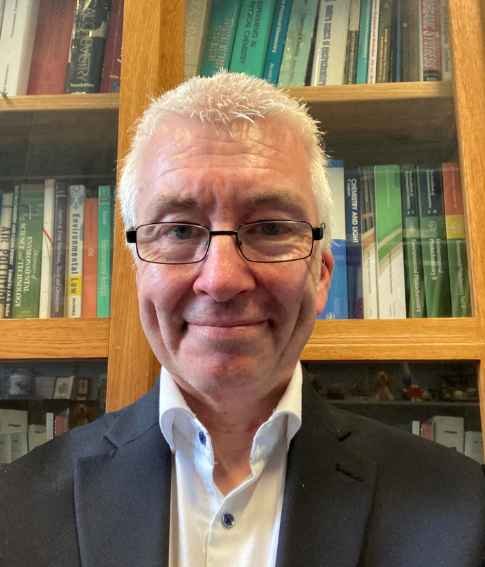 Photo of Peter, smiling and wearing a blazer standing in front of his book shelf.