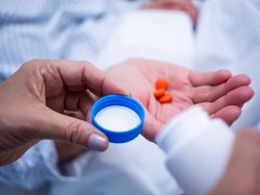 A close up two people's hands, one holding an open pill bottle the other holding orange pills in their right hand.