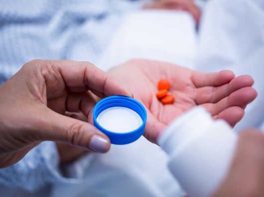 A close up two people's hands, one holding an open pill bottle the other holding orange pills in their right hand.