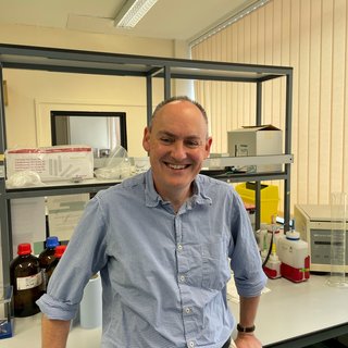 A picture of a man stood in the lab smiling wearing a blue and white checked shirt.