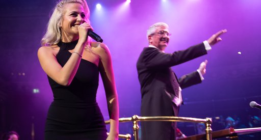 A lady holding a microphone and singing at a concert hall with people cheering in the background.