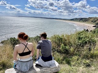 Two young people sitting on a bench, viewed from behind. They're looking out to sea on a sunny day.