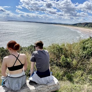 Two young people sitting on a bench, viewed from behind. They're looking out to sea on a sunny day.