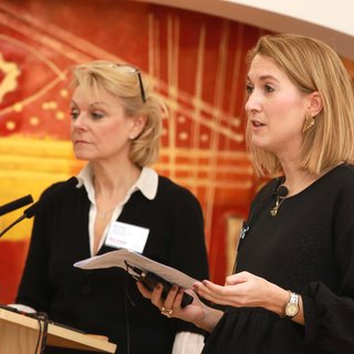 Two women stand at a lectern. One of the women holds a piece of paper.