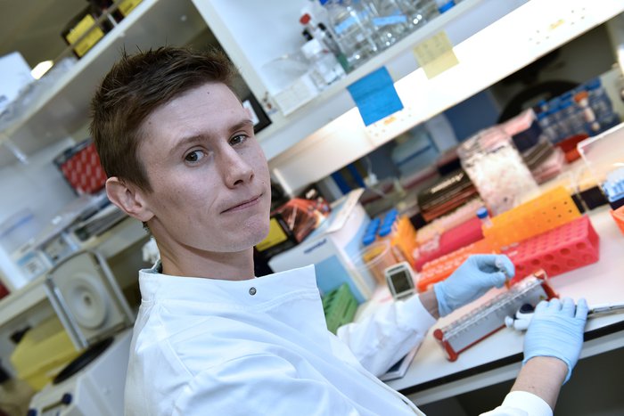 Dan Coleman, from the University of Birmingham, stands in a laboratory holding a test tube.