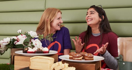 Two people wearing Blood Cancer UK t-shirts, smiling behind a beautiful spread of cakes and cupcakes.