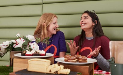 Two members of Blood Cancer UK staff wearing branded t-shirts whilst laughing at a table covered in plates of cakes.