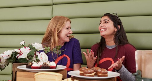 Two members of Blood Cancer UK staff wearing branded t-shirts whilst laughing at a table covered in plates of cakes.