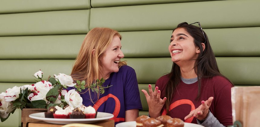 Two members of Blood Cancer UK staff wearing branded t-shirts whilst laughing at a table covered in plates of cakes.