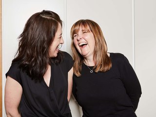 Two people standing side-by-side looking happy and laughing together against a wall in a house.