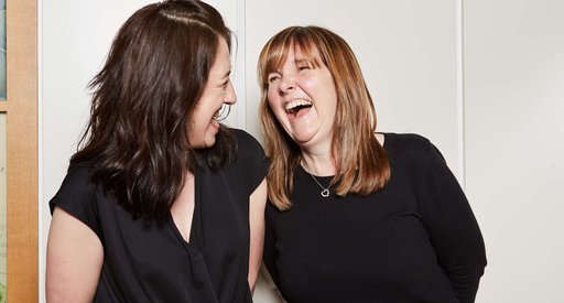 Two people standing side-by-side looking happy and laughing together against a wall in a house.