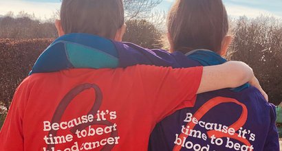 Two supporters hug, wearing Blood Cancer UK T-shirts, with the slogan "Because it's time to beat blood cancer"