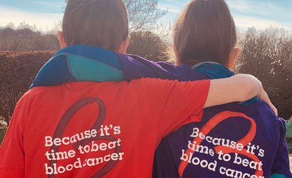 Two supporters hug, wearing Blood Cancer UK T-shirts, with the slogan "Because it's time to beat blood cancer"