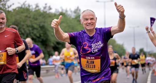 An older man in a purple Blood Cancer UK t-shirt holds his thumbs up as he runs a marathon.