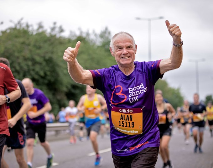 An older man in a purple Blood Cancer UK t-shirt holds his thumbs up as he runs a marathon.