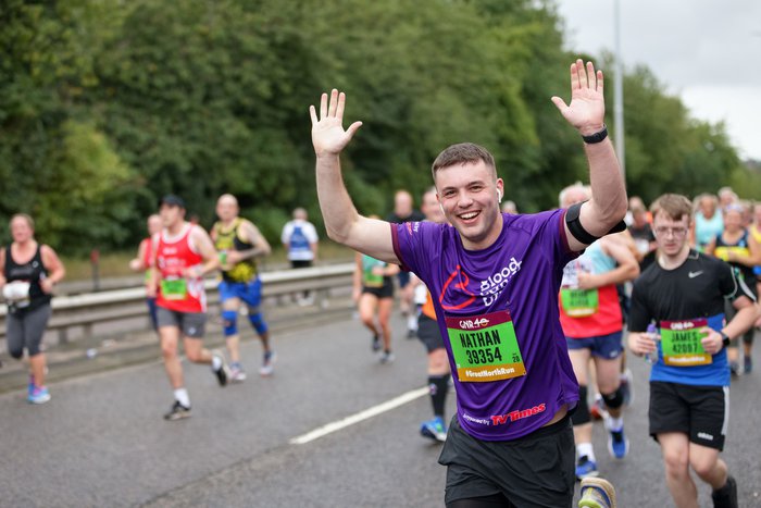 A man cheers and raises his hands in the air as he runs through the city for the Great North Run, wearing a purple Blood Cancer UK running vest