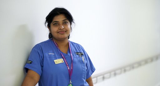 A nurse wearing blue scrubs, smiling in the hallway of a hospital.