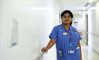 A nurse in a hallway of a hospital, leaning against an arm rail, smiling in her blue scrubs.