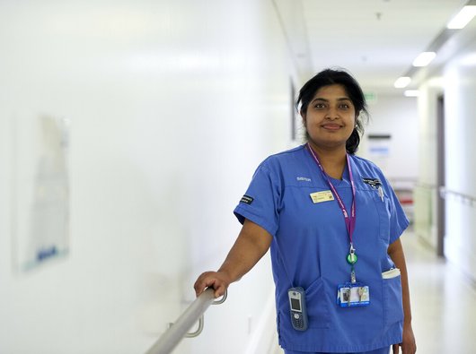 A nurse in a hallway of a hospital, leaning against an arm rail, smiling in her blue scrubs.