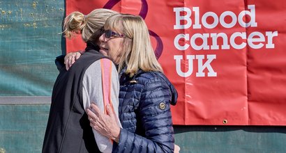 Two women hug each other in front of a Blood Cancer UK banner