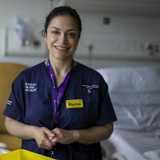 A close up of a healthcare professional wearing blue scrubs, smiling at the camera with hospital equipment out of focus in the background.