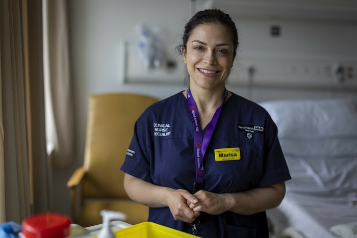 A close up of a healthcare professional wearing blue scrubs, smiling at the camera with hospital equipment out of focus in the background.